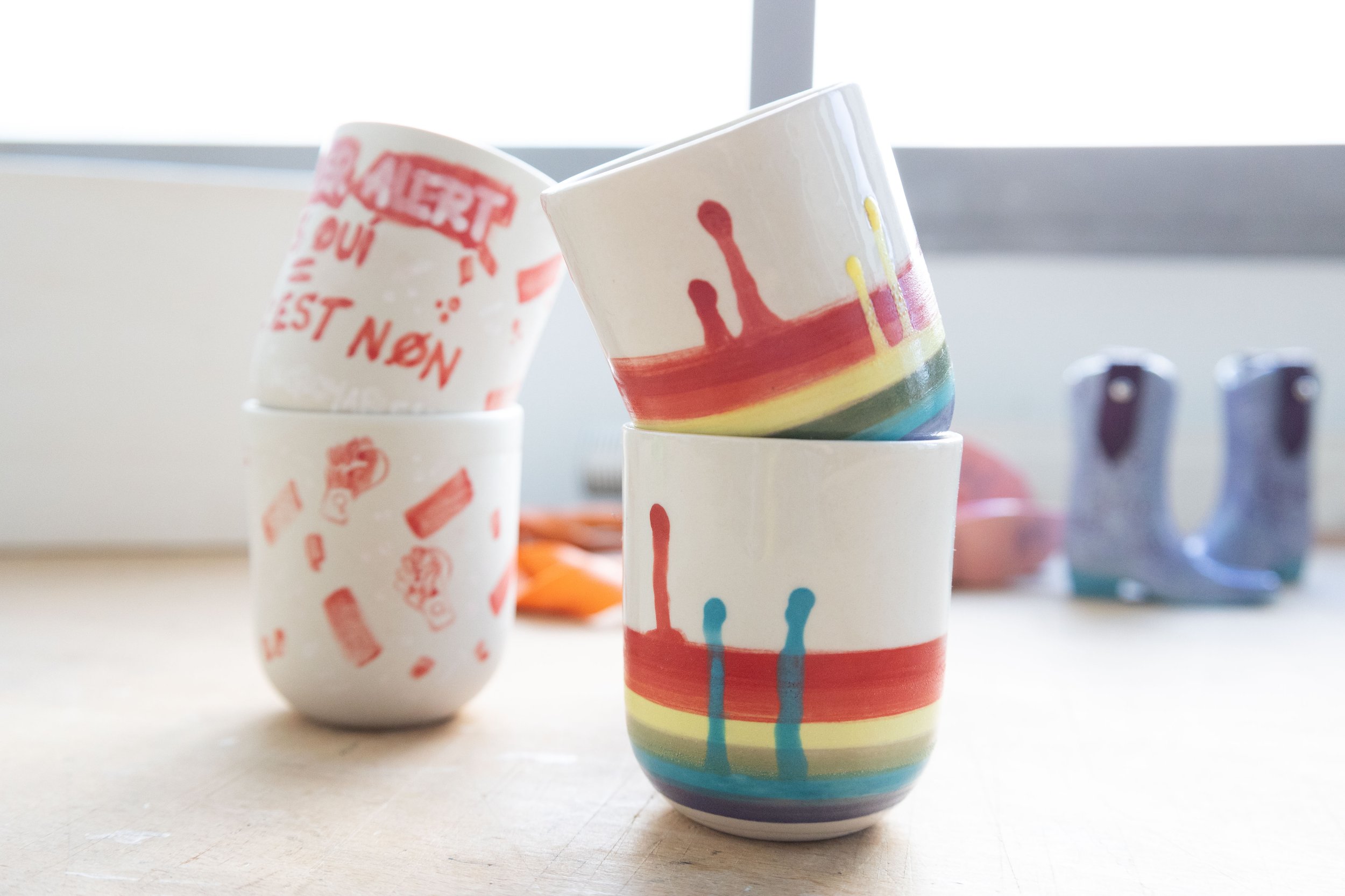 Tasses en céramique empilées, avec des motifs peints à la main, dont un arc-en-ciel et des dessins rouges sur fond blanc, posées sur une table en bois, avec des objets flous à l'arrière-plan.